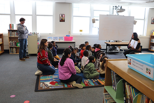 Students seating down, listening to a teacher reading a book